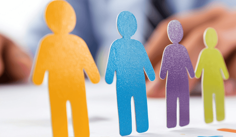Colorful cutout figures representing different teams or portfolio segments stand on a desk covered with bar graphs, with a blurred businessperson in the background reaching toward them—symbolizing portfolio oversight, segmentation, or organizational analysis.